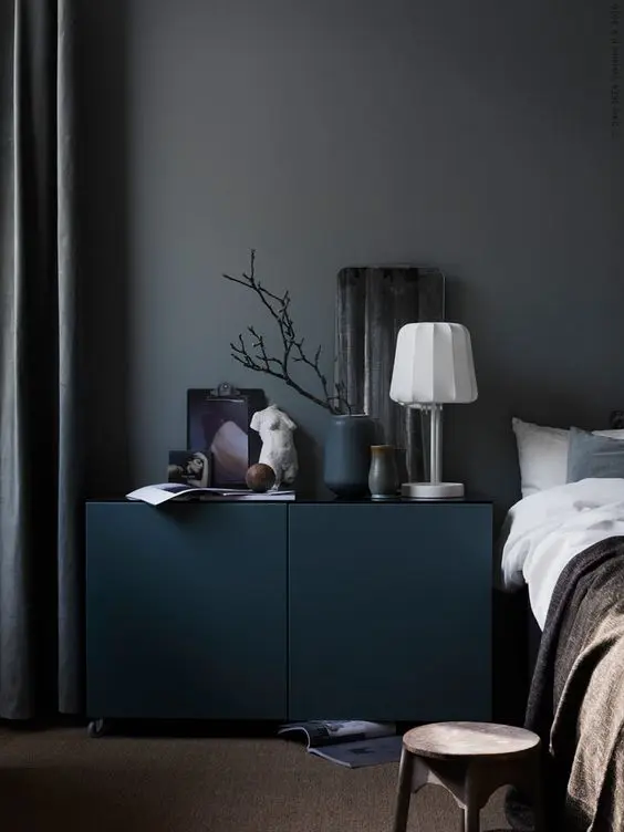 a moody and cool bedroom with grey walls, a teal cabinet, a bed with neutral bedding, some decor and a table lamp is a chic and lovely space