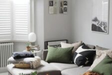 a Nordic living room with light grey walls,a  bay window with shutters, a grey sectional with green and other pillows, a coffee table and a woven pendant lamp