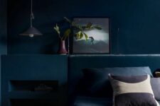 a beautiful dark bedroom with navy walls and a built-in matching niche, a navy upholstered bed with navy bedding, some art and a pendant lamp