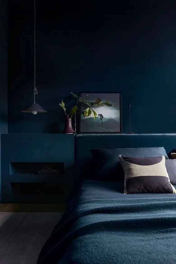 A beautiful dark bedroom with navy walls and a built in matching niche, a navy upholstered bed with navy bedding, some art and a pendant lamp