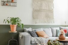 a boho living room with a color block accent wall in green and white, with a light grey sofa with pillows, a macrame, potted greenery
