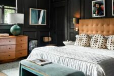 a chic moody bedroom with black molded walls, an amber leather bed with neutral bedding, a green upholstered bench and a stained dresser