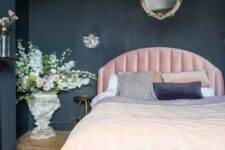 a chic moody bedroom with black walls and a chest for storage, a light pink bed, refined lighting and blooms
