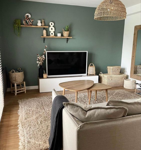 a cute Scandinavian living room with a green accent wall, a TV unit and a TV, some shelves, baskets and a greige sofa plus coffee tables