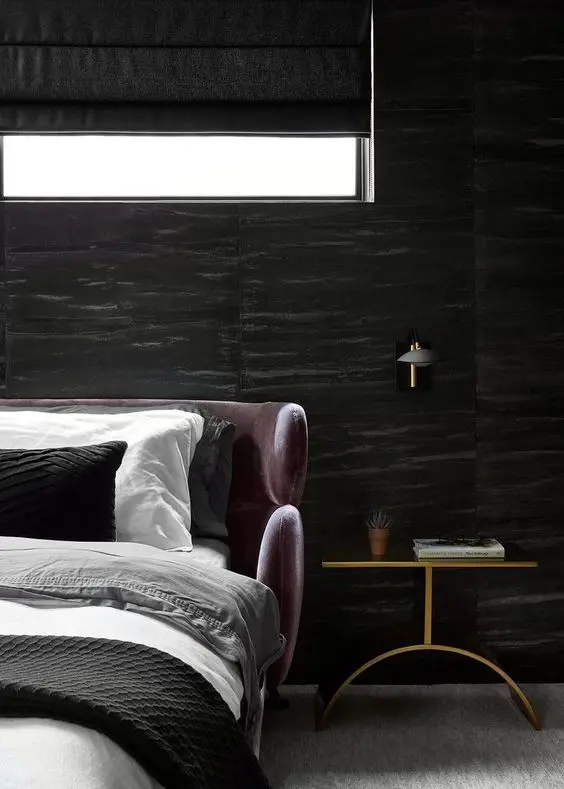 a gorgeous dark bedroom with black fabric upholstered walls, a mauve upholstered bed with neutral bedding, a gold nightstand and a sconce