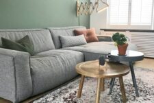 a laconic living room with a green accent wall, a low grey sectional, a couple of coffee tables, a catchy wall lamp and a printed rug