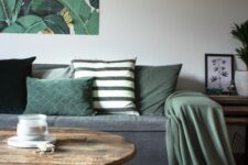 a lovely living room with a graphite grey sofa and rug, green pillows and a blanket, a bold artwork, a coffee table and a pendant lamp