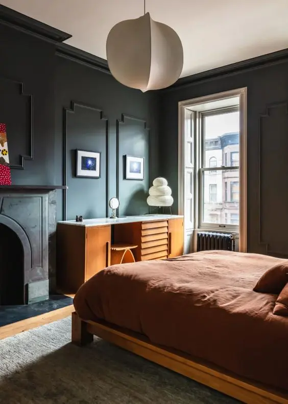 a lovely moody bedroom with black trim walls, a non-working fireplace, a stained bed with rust bedding, a stained desk