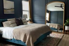 a moody and peaceful bedroom with soot walls, a blue upholstered bed with neutral bedding, a large arched floor mirror and a printed rug