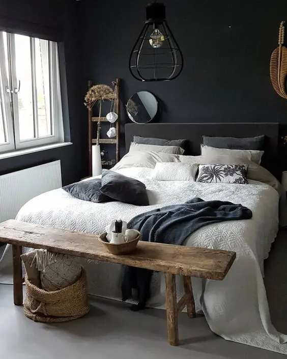 A moody bedroom with black walls, a grey upholstered bed, neutral bedding, a rough stained bench, baskets and a ladder plus some wabi sabi decor