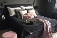 a moody bedroom with grey walls, a black bed, nightstand and a pendant lamp, pink and black bedding and a pink ottoman