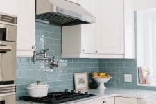 a neutral farmhouse kitchen with shaker cabinets, a blue subway tile backsplash and a metal hood, white stone countertops