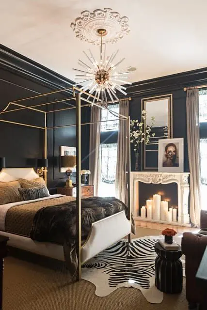 a refined moody bedroom with black walls, a faux fireplace with pillar candles, some art, a canopy bed, a burst chandelier and a sofa