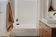 a small and cozy bathroon done with white subway and black hex tiles, a wooden vanity, neutral linens and a wooden stool