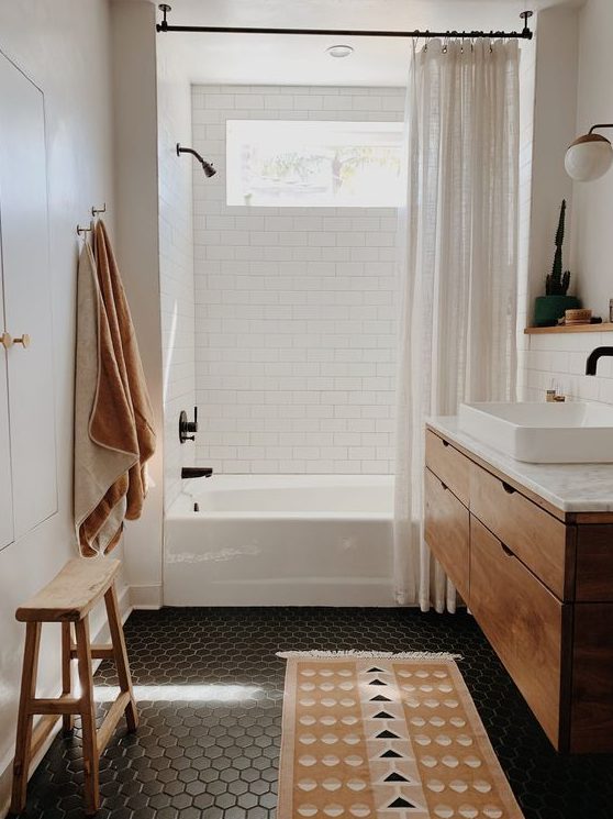 a small and cozy bathroon done with white subway and black hex tiles, a wooden vanity, neutral linens and a wooden stool