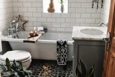 a small boho bathroom with a mosaic tile floor, grey and white tile walls, a tub, potted greenery and baskets for storage