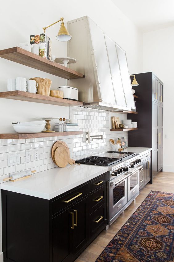 a stylish black farmhouse kitchen with white stone countertops, a whiet subway tile backsplash, open shelves and a large metal hood