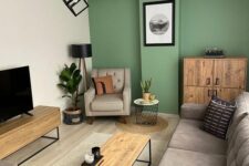 a stylish living room with a green accent wall, a large greige sectional, a TV on a TV unit, a black coffee table, a chair and a storage unit