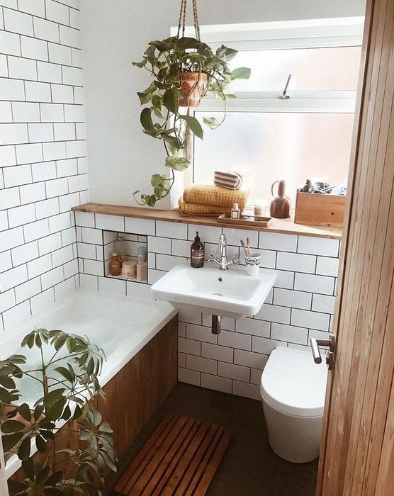 A tiny mid century modern bathroom with white subway tiles, a bathtub clad with woode, potted greenery and a window