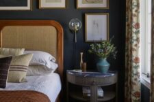 a vintage moody bedroom with soot walls, a bed with a cane headboard, a fluted nightstand, a gallery wall and printed textiles