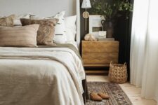 a welcoming moody bedroom with a soot accent wall, a neutral upholstered bed with neutral bedding, a printed rug, pendant plants and an inlay nightstand