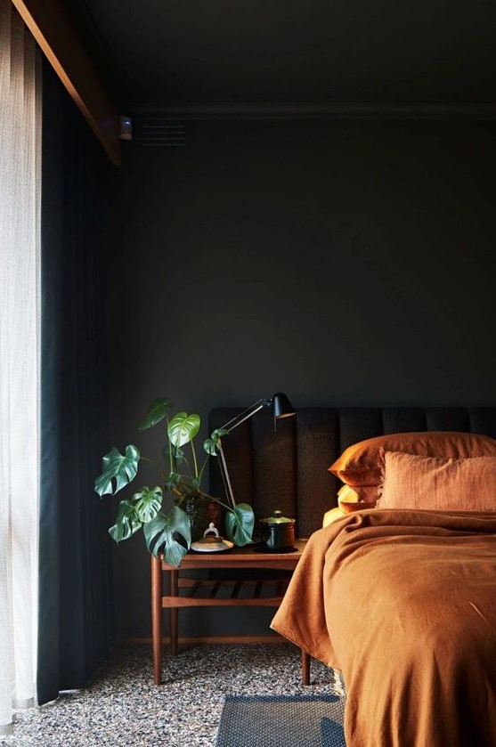 A welcoming moody bedroom with black walls and a bed, with wooden nightstands, rust colored bedding and black curtains