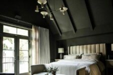 an attic bedroom with black walls and a ceiling, a neutral upholstered bed with neutral bedding, a neutral bench, a black chair and a chandelier