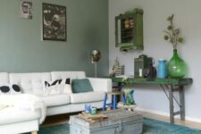 an eclectic living room with a grey and green wall, a white sectional with pillows, a green shabby chic console table and a suitcase as a table