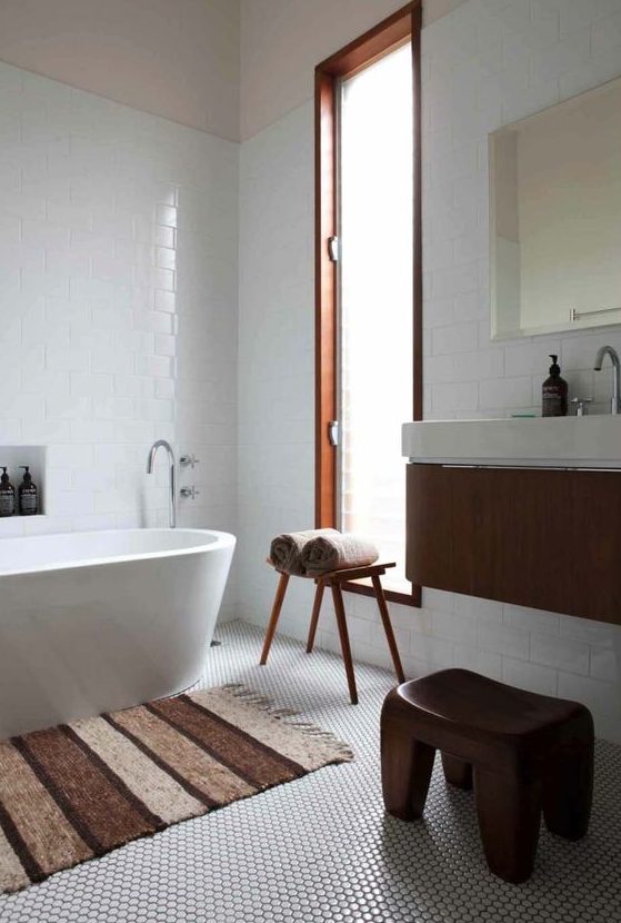 an ethereal mid-century modern bathroom with white penny and subway tiles, an oval tub, a floating vanity and a wooden stool