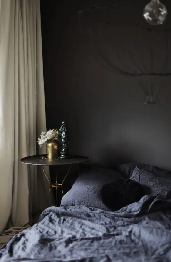 a dark moody bedroom with a black accent wall, a bed with graphite bedding, a nightstand with gold legs and neutral curtains