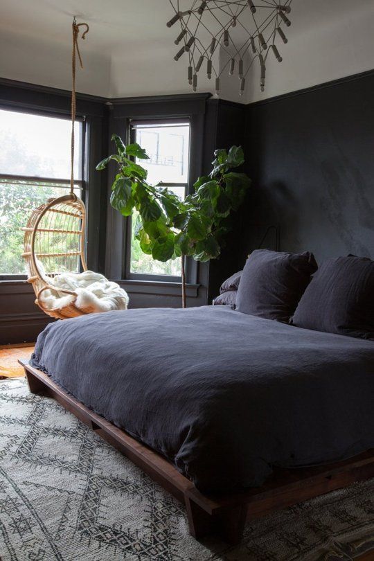 a dark boho bedroom with a reclaimed wood bed, a hanging chair by the window and a potted tree, a catchy chandelier