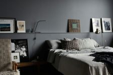 a moody bedroom with graphite grey walls, a bed with greyish bedding, a ledge with artwork and a woven chair plus some pillows