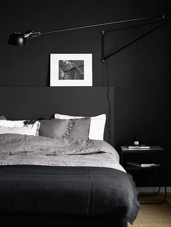 a minimalist bedroom design with matte black walls, a black bed with monochromatic bedding, a nightstand and a black lamp plus an artwork