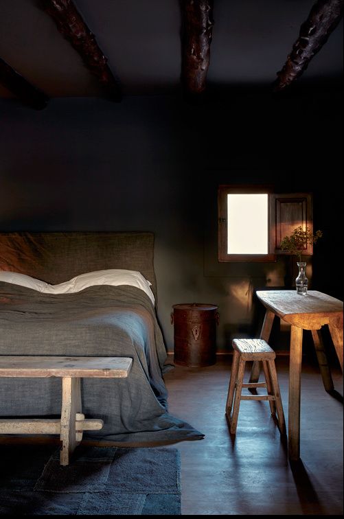 A rustic moody bedroom decor with dark walls, dark stained wooden beams, an upholstered bed and rough wooden furniture