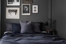 a tiny moody bedroom with grey walls, a bed with navy and black bedding, a gallery wall and a table lamp is cool and chic