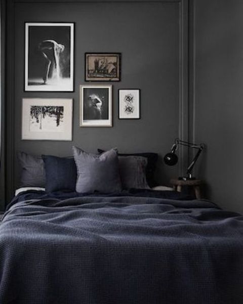 a tiny moody bedroom with grey walls, a bed with navy and black bedding, a gallery wall and a table lamp is cool and chic