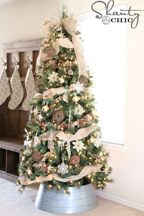 a rustic tree decorated with burlap, gold and white ornaments, snowflakes and lights, pinecones and ribbons in a galvanized bucket