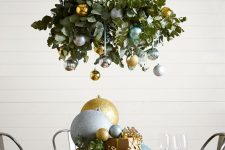 a chic holiday chandelier of eucalyptus, silver, gold and green ornaments and matching oversized ones on the table
