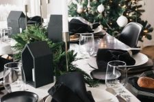 a modern black and white Christmas tablescape with a printed runner, black napkins, black house-shaped candleholders and evergreens and white candles