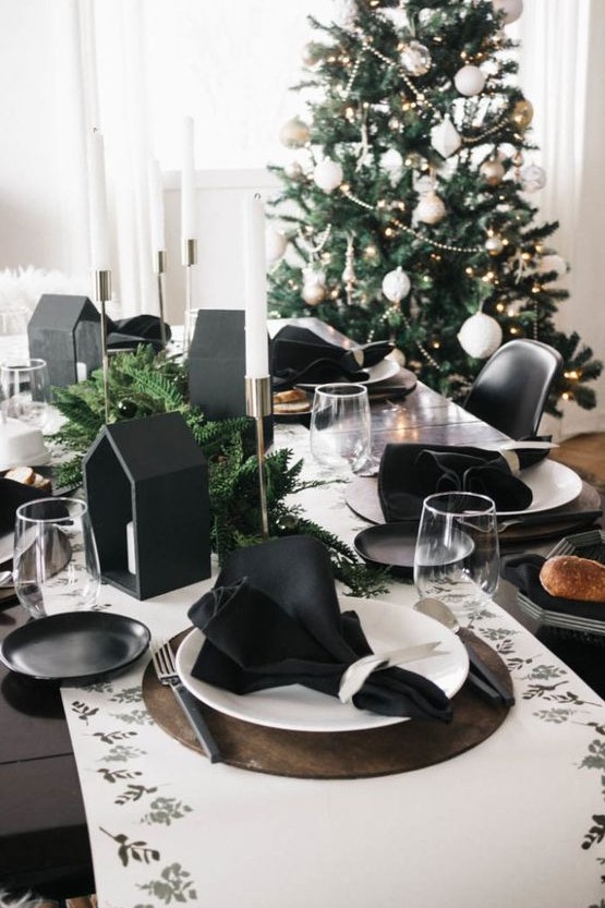 A modern black and white Christmas tablescape with a printed runner, black napkins, black house shaped candleholders and evergreens and white candles