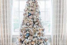 a pretty flocked Christmas tree with brown ribbons, gold and navy ornaments, lights and frozen branches on top