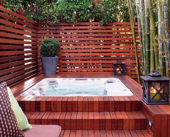 a wood clad jacuzzi with a plank wall around and lanterns and greenery in pots