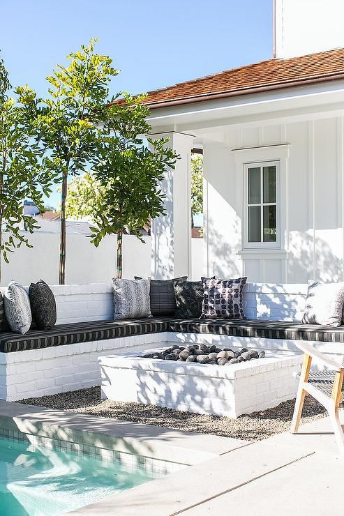 an L shaped white brick bench lined with black and white cushions facing a white brick fire pit and a ground pool