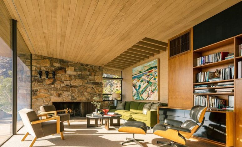 The living room features a natural wood ceiling with beams, s tone clad wall with a fireplace and a fine selection of comfy mid-century furniture