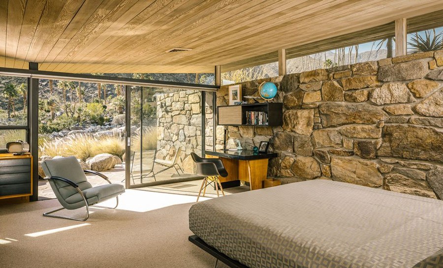The master bedroom shows the same ceiling and a stone clad wall, comfy furniture in warm shades and an entrance to the terrace, this glazed wall lets enjoying the views