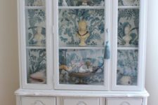 23 a white vintage cabinet with blue and white wallpaper inside for displaying souvenirs