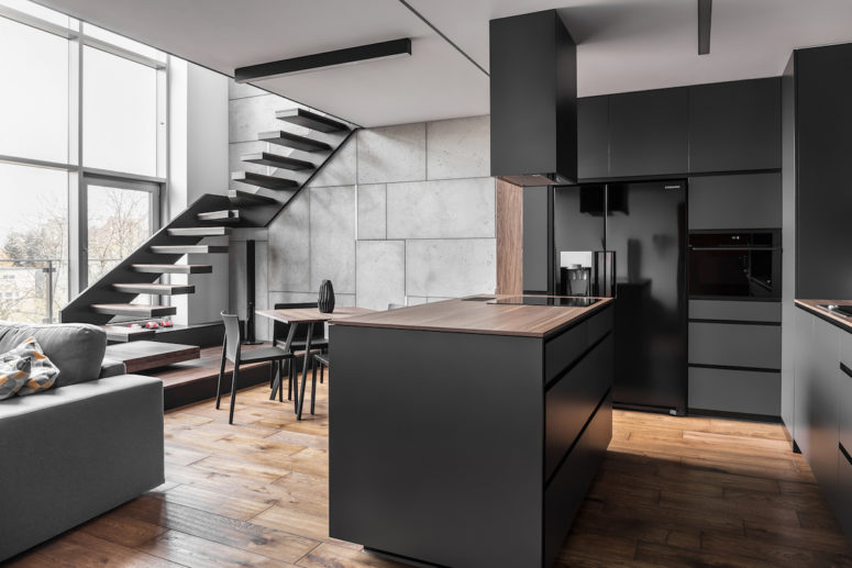 The kitchen is done in matte black and grey, large windows fill the open layout with light, and a linear staircase leaves some space for an eating zone