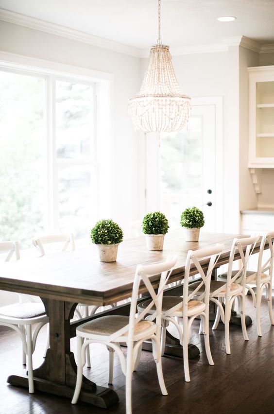 a cottage dining room with white chairs and a wooden trestle table, a glam chandelier to add style to the space