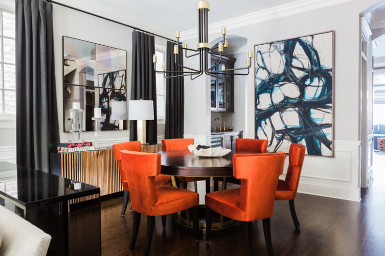 The dining area is bold and glam, there's a large artwork, a mirror, a brass and black chandelier and super colorful burnt orange chairs