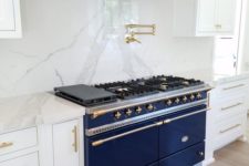 16 a white kitchen is spruced up with marble, a brass hood and a large cobalt blue cooker